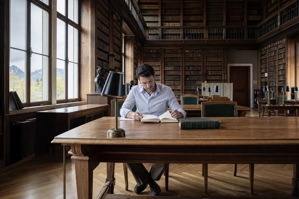 Mann liest an einem langen Tisch in einer historischen Bibliothek mit hohen Bücherregalen